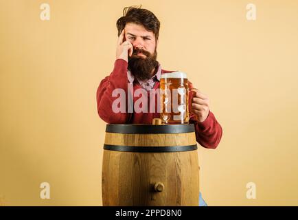 Tempo di birra. Macchina da caffè premurosa con tazza di birra su barile di legno. Uomo serio barbuto con birra artigianale. Vacanze, bevande, alcol e tempo libero. Barbuto Foto Stock