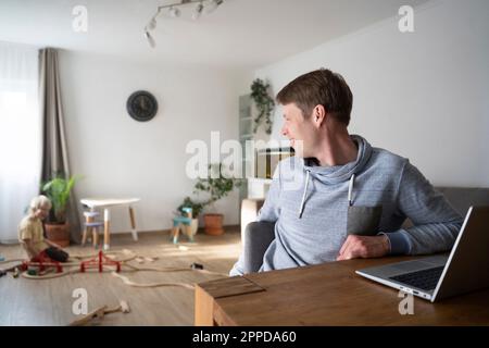 Felice freelance seduto alla scrivania con il figlio che gioca in background Foto Stock
