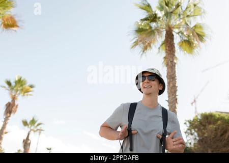 Uomo felice che indossa il cappello e con lo zaino in piedi davanti alla palma Foto Stock