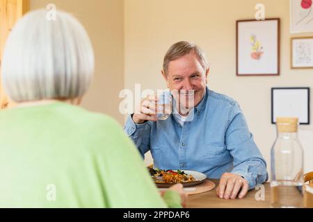 Uomo anziano sorridente con un bicchiere d'acqua gustando il pranzo a casa Foto Stock