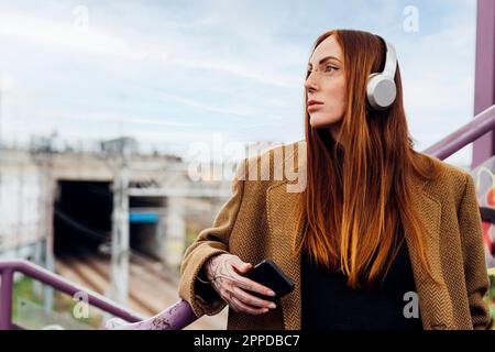 Donna dalla testa rossa attenta che indossa cuffie wireless per ascoltare la musica Foto Stock