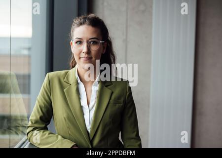 Donna d'affari giovane e sicura che indossa occhiali in piedi dalla finestra Foto Stock