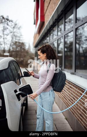 Donna sorridente che usa lo smartphone e ricarica l'auto elettrica Foto Stock