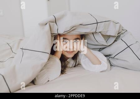 Ragazza simpatica e sorridente sdraiata sotto la coperta sul letto Foto Stock
