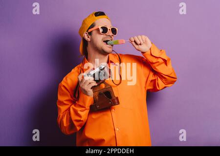 Uomo elegante in camicia arancione con macchina fotografica e gelato su sfondo viola Foto Stock