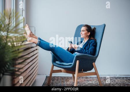 Felice giovane donna d'affari che tiene lo smartphone seduto in poltrona Foto Stock