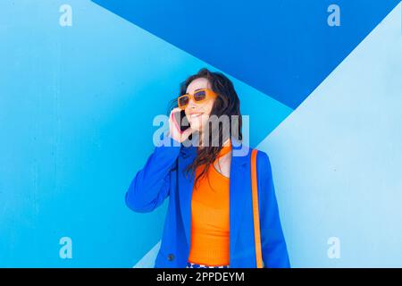 Donna felice alla moda che indossa occhiali da sole che parlano sullo smartphone di fronte alla parete blu Foto Stock