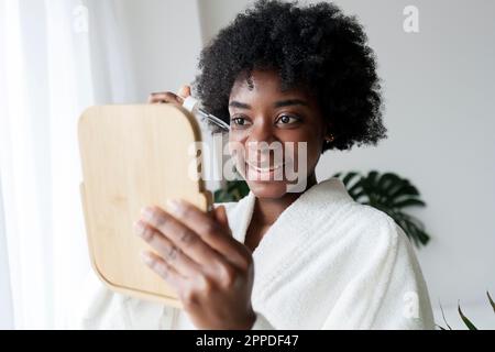 Giovane donna sorridente che applica siero sul viso a casa Foto Stock