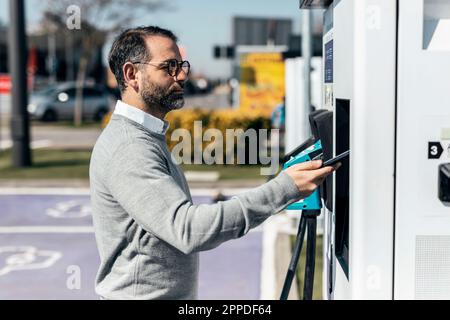 Uomo che paga tramite smartphone alla stazione di ricarica per auto Foto Stock