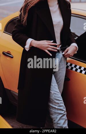 Figura femminile in jeans denim blu, maglione bianco e lungo cappotto nero che cammina contro il taxi giallo. Stile urbano autunnale Foto Stock