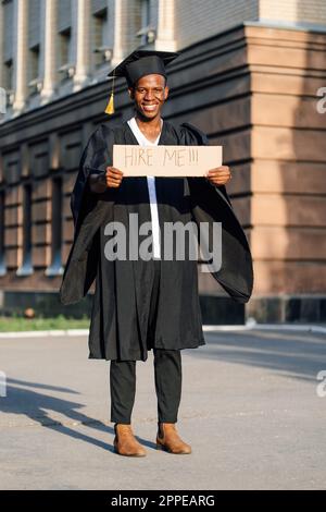 Gioire ragazzo nero in piedi con poster di cartone sulla strada in giornata di sole in cerca di lavoro. Università o università laureando studente in abito e tappo Foto Stock