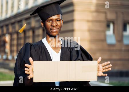 Povero ragazzo nero in piedi con cartoncino cartello sulla strada in cerca di lavoro. Studente universitario o universitario che si laurea indossando abito da laurea e berretto Foto Stock