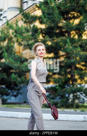 Ritratto lunghezza intera di bella donna che cammina all'aperto tra abeti verdi con borsa a spalla in pelle borgogna in mano. Di mezza età, 40s donna felice Foto Stock