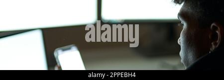 Primo piano di un giovane ragazzo occupato seduto al buio di fronte a molti monitor di computer e utilizzando smartphone mentre lavorava da solo fino a tarda notte Foto Stock