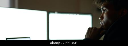 Lavoro tutta la notte. Giovane ragazzo concentrato seduto in una stanza buia di fronte a molti monitor di computer mentre lavorava da solo a tarda notte Foto Stock