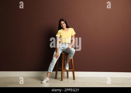 Bella giovane donna seduta su sgabello vicino al muro marrone Foto Stock