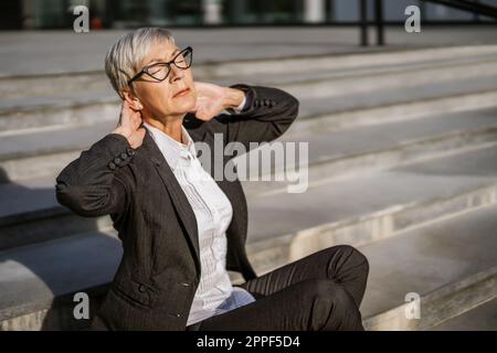 Donna d'affari matura seduta di fronte alla costruzione di società. È stanca e ha dolore al collo. Foto Stock