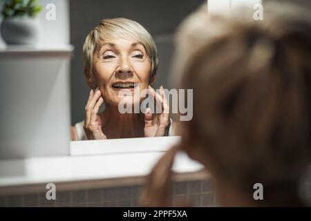 Donna matura che guarda il suo volto in bagno. Sta esaminando le rughe sulla sua pelle. Foto Stock
