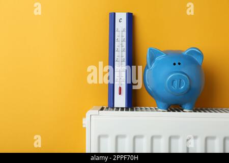 Piggy bank e termometro sul radiatore di riscaldamento su sfondo arancione, spazio per testo Foto Stock