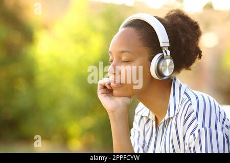 Donna nera rilassata che indossa cuffie ascoltare musica riposando in un parco Foto Stock