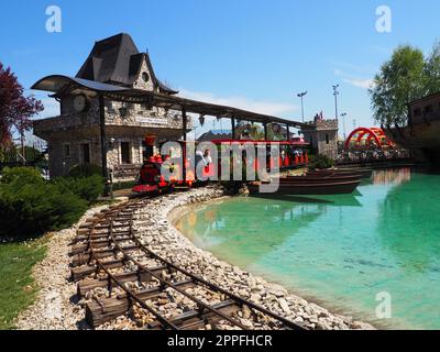 Stanisici, Bijelina, BiH, 25 aprile 2021. Una locomotiva a vapore per bambini con persone e bambini guida i passeggeri. La locomotiva e i carri sono dipinti di rosso. Parco divertimenti estivo Foto Stock