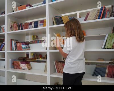 Sremska Mitrovica, Serbia 15 maggio 2021. Una bambina di 7 anni con una maglietta bianca si trova vicino alla libreria della biblioteca e legge un libro. Istruzione scolastica. Amore per la letteratura e la lettura Foto Stock