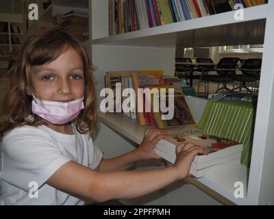 Sremska Mitrovica, Serbia 15 maggio 2021. Una bambina di 7 anni che indossa una maschera e una maglietta bianca si trova vicino a una libreria e tocca i libri con le mani. Istruzione scolastica, letteratura e lettura Foto Stock