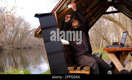 L'uomo lavora con la ricarica di un computer portatile da un pannello solare in alclove vicino al fiume. Energia pulita da utilizzare in campeggio. Lavora con gadget all'aperto con pannelli solari portatili. Foto Stock