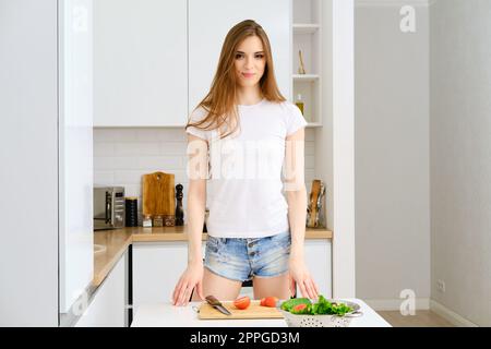 Giovane bella casalinga con capelli lunghi sta andando a preparare insalata di verdure sana in moderna cucina bianca a casa Foto Stock