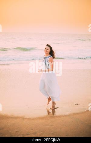 Goa, India. Giovane donna caucasica in abito bianco camminando lungo Seashore, guardando la macchina fotografica e sorridendo in estate luce del sole Foto Stock