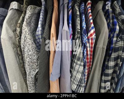 Vestiti appesi agli appendiabiti. Assortimento di articoli di seconda mano. Camicie da uomo appese a un rack, pronte per la vendita ai clienti. Risparmiare denaro in crisi. Shopping economico. Camicie a scacchi, a righe, semplici da uomo Foto Stock