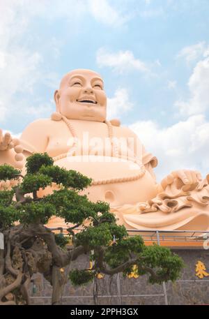 Statua di buddha ridente al tempio di Vinh Trang, vicino a My Tho, Vietnam. Vista ad angolo basso con albero bonsai in primo piano. Foto Stock