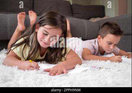 Fratelli che giocano insieme a casa. bambino e ragazza sdraiati sul tappeto e disegnati su fogli di carta bianchi con pastelli colorati Foto Stock