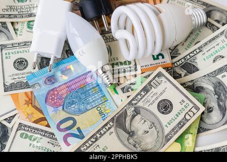 Il concetto di crisi energetica, i prezzi aumentano. Banconote in euro e in dollari, monete sullo sfondo di una lampada, spina, cavo. Tempo fiscale, pagamento fattura, calcolatore per il conteggio. Foto Stock