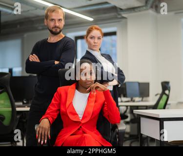 Donna caucasica dai capelli rossi, uomo caucasico bearded in piedi dietro una giovane donna afroamericana seduta in ufficio. Foto Stock