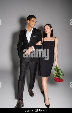 intera lunghezza di felice uomo afroamericano ed elegante donna asiatica con rose rosse che camminano su sfondo grigio, immagine di scorta Foto Stock
