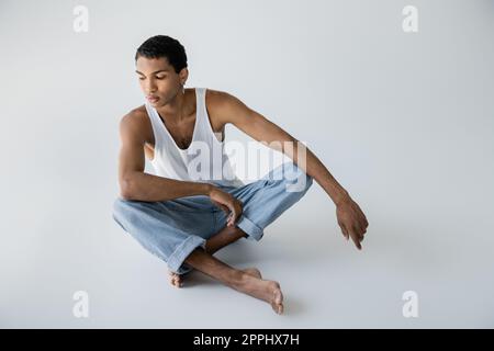 uomo afro-americano a piedi nudi in jeans seduti con gambe incrociate su sfondo grigio, immagine stock Foto Stock