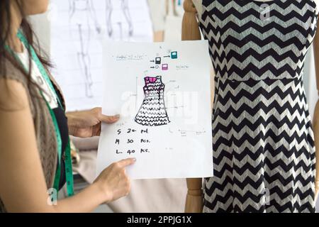 Giovane proprietario di affari di disegno di moda che controlla gli ordini dalla nota sul modello per il panno, comparando con il vestito sul manichino. Foto Stock