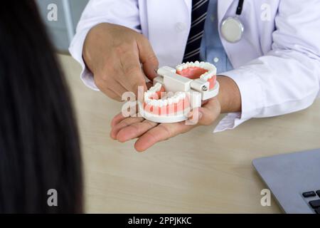 Dentista in abito bianco e stetoscopio che punta il dito sul modello di dente, spiega ai giovani pazienti l'ortodonzia e l'apparecchio. Aiuta a ridurre il dolore gengivale. Concetto di sanità e medicina. Primo piano Foto Stock