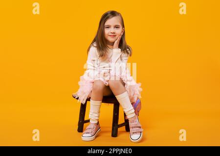 Ragazza giovane in gonna tulle e pullover bianco seduta su sgabello in legno su sfondo giallo brillante Foto Stock