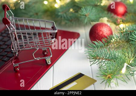 Carrello per la spesa su tastiera di computer portatile, carta di credito, rami dell'albero di Natale con lampadine e palline su sfondo bianco Foto Stock