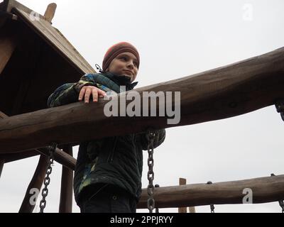 ragazzo di 8 anni al parco giochi. Il bambino è vestito con abiti caldi e stagionali - un cappello marrone e una giacca verde. Area di arrampicata in legno per bambini. Ragazzo che guarda lontano Foto Stock