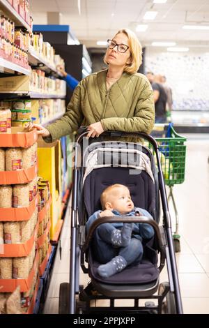 La madre vestita Casualy sceglie prodotti in scatola nel reparto del supermercato, con il bambino in passeggino. Foto Stock