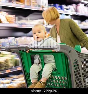 Casualy ha vestito la madre che sceglie i prodotti nel reparto del supermercato, con il suo bambino neonato nel carrello della spesa. Foto Stock