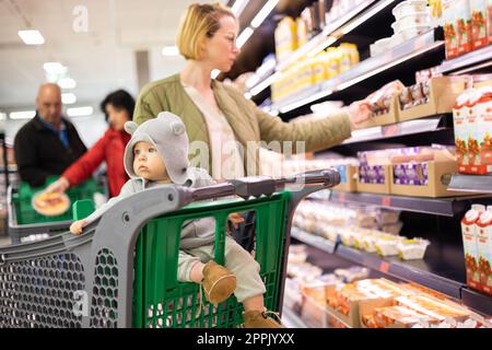 Casualy ha vestito la madre che sceglie i prodotti nel reparto del supermercato, con il suo bambino neonato nel carrello della spesa. Foto Stock