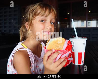 Anapa Russia 23 agosto 2021 Una bella ragazza di 7 anni sta mangiando al ristorante KFC gustando il cibo e sorridendo. Bambino caucasico che tiene un hamburger. Coca cola in una tazza di cartone con coperchio e cannuccia Foto Stock