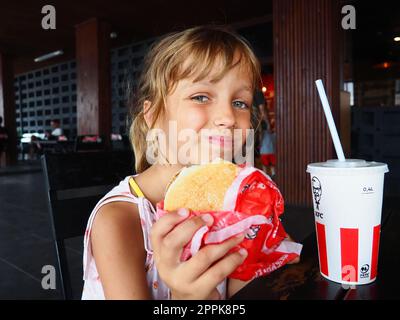 Anapa Russia 23 agosto 2021 Una bella ragazza di 7 anni sta mangiando al ristorante KFC gustando il cibo e sorridendo. Bambino caucasico che tiene un hamburger. Coca cola in una tazza di cartone con coperchio e cannuccia Foto Stock