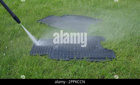 Lavare i tappetini neri in gomma per auto con l'idropulitrice manuale sul prato verde. Spruzzi d'acqua sul manto erboso Foto Stock