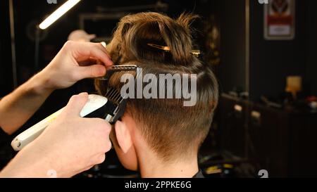 acconciatura e taglio di capelli da uomo con regolacapelli in un barbiere o in un parrucchiere. Servizio di parrucchiere in un moderno barbiere con illuminazione a chiave scura e calda vista sul retro Foto Stock