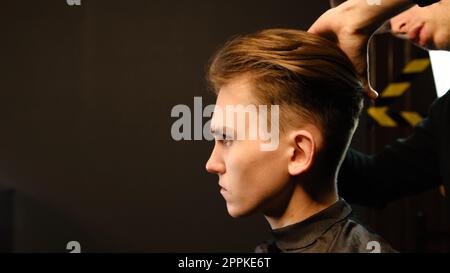 Barbiere. L'uomo in poltrona da barbiere, parrucchiere, prepara i capelli con il pettine per tagliarli e modellarli. Concept Barbershop. vista laterale con illuminazione principale scura e luce calda Foto Stock